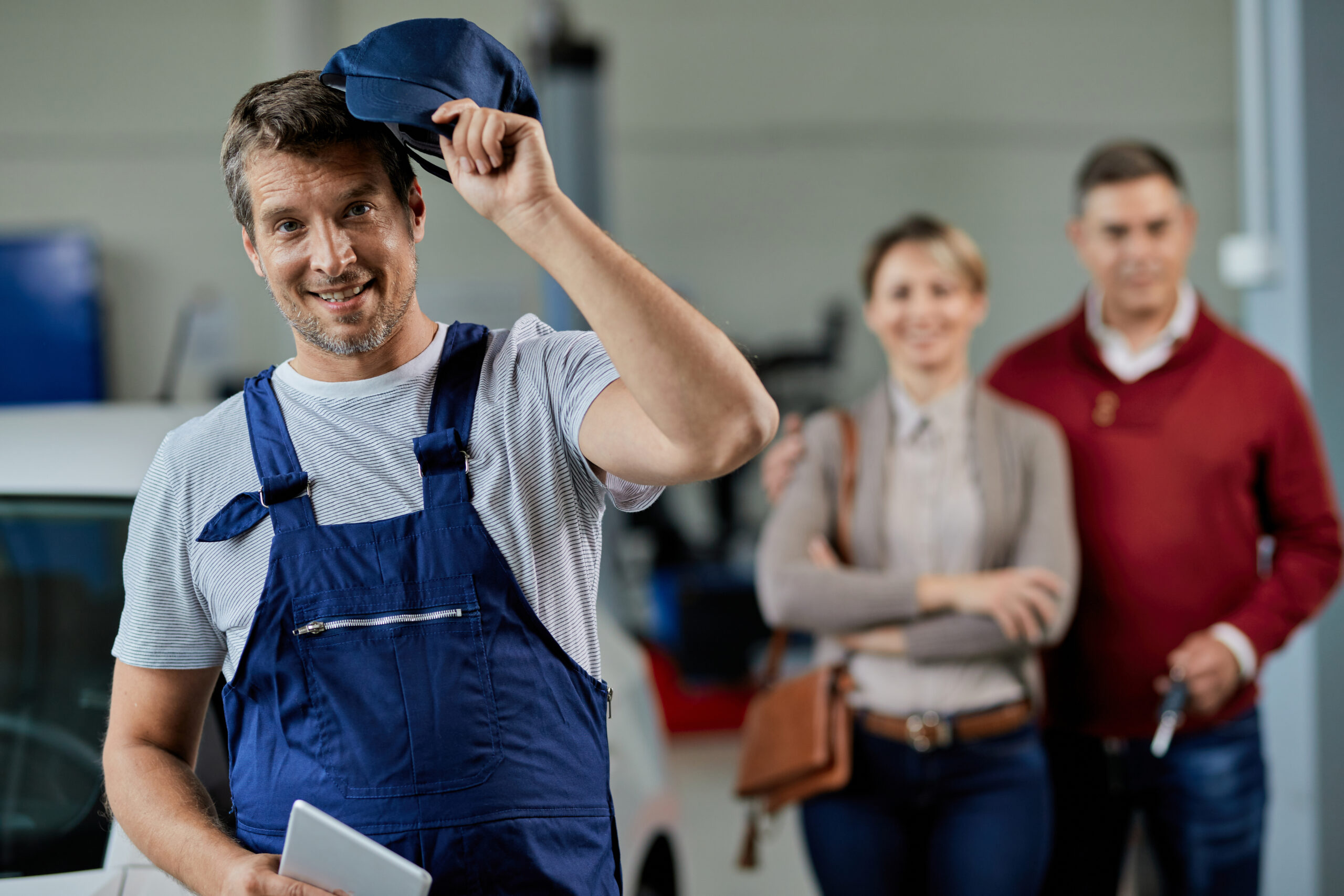 Satisfaction of our customers is our top priority! Happy auto mechanic taking his hat of while standing in a workshop and looking at camera. His customers are in the background.